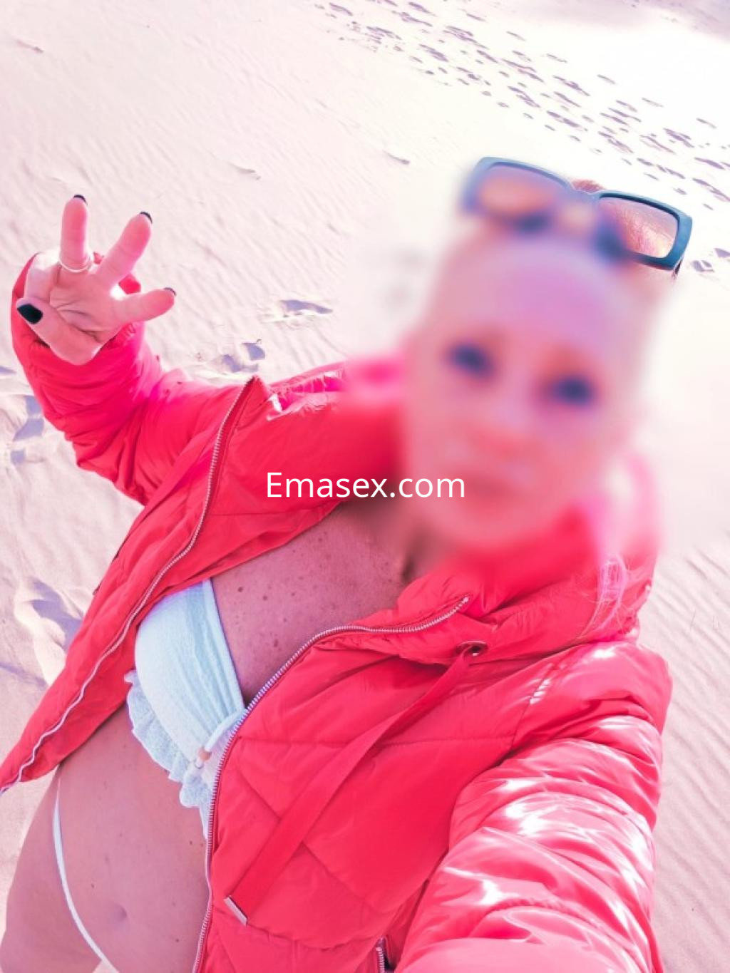 Escort en Lugo posando en la playa con una chaqueta roja y bikini claro, mostrando un gesto de paz.