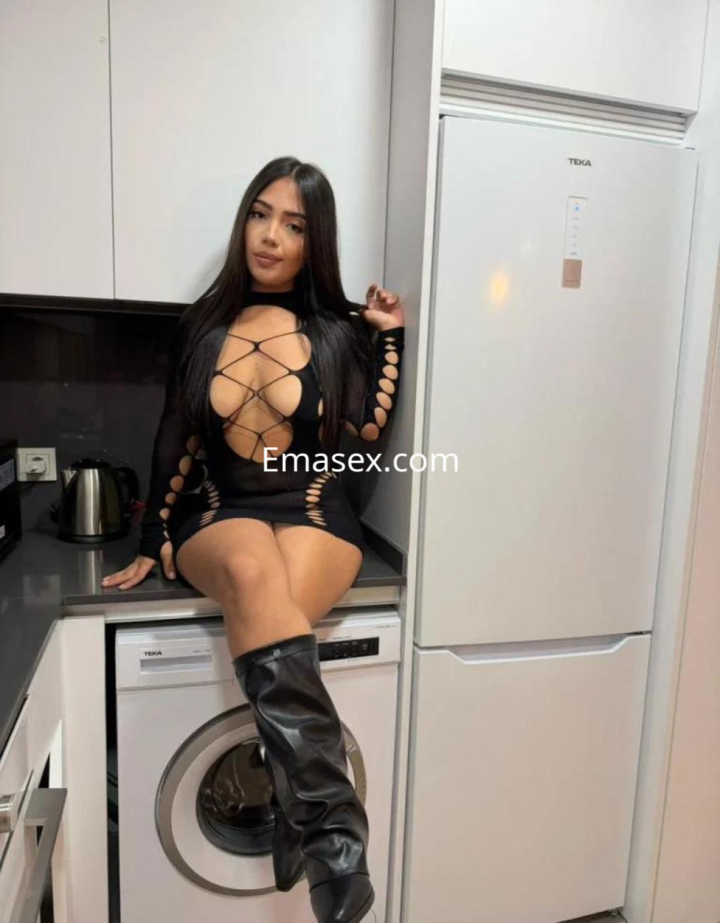 Escort en Málaga posando en una cocina moderna, vistiendo un vestido negro ajustado con detalles cut-out y botas altas.