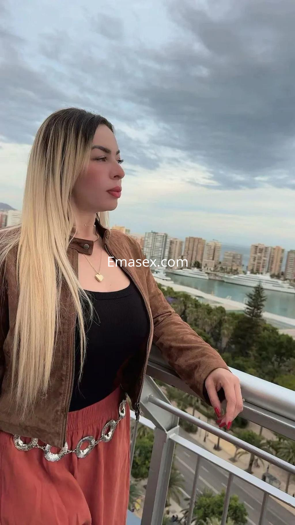 Escort de Málaga con cabello largo y rubio, vistiendo un top negro y una falda larga de color terracota, posando en un balcón.