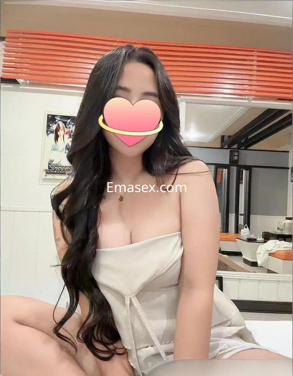 Puta con cabello largo y oscuro, sentada en una habitación con paredes claras, vistiendo una blusa blanca.
