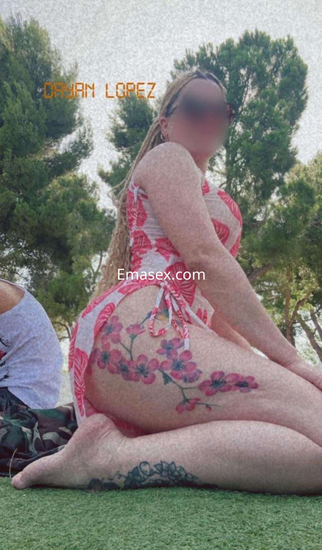 Escort en Toledo con vestido de flores, mostrando un tatuaje en la pierna y posando en un entorno al aire libre.