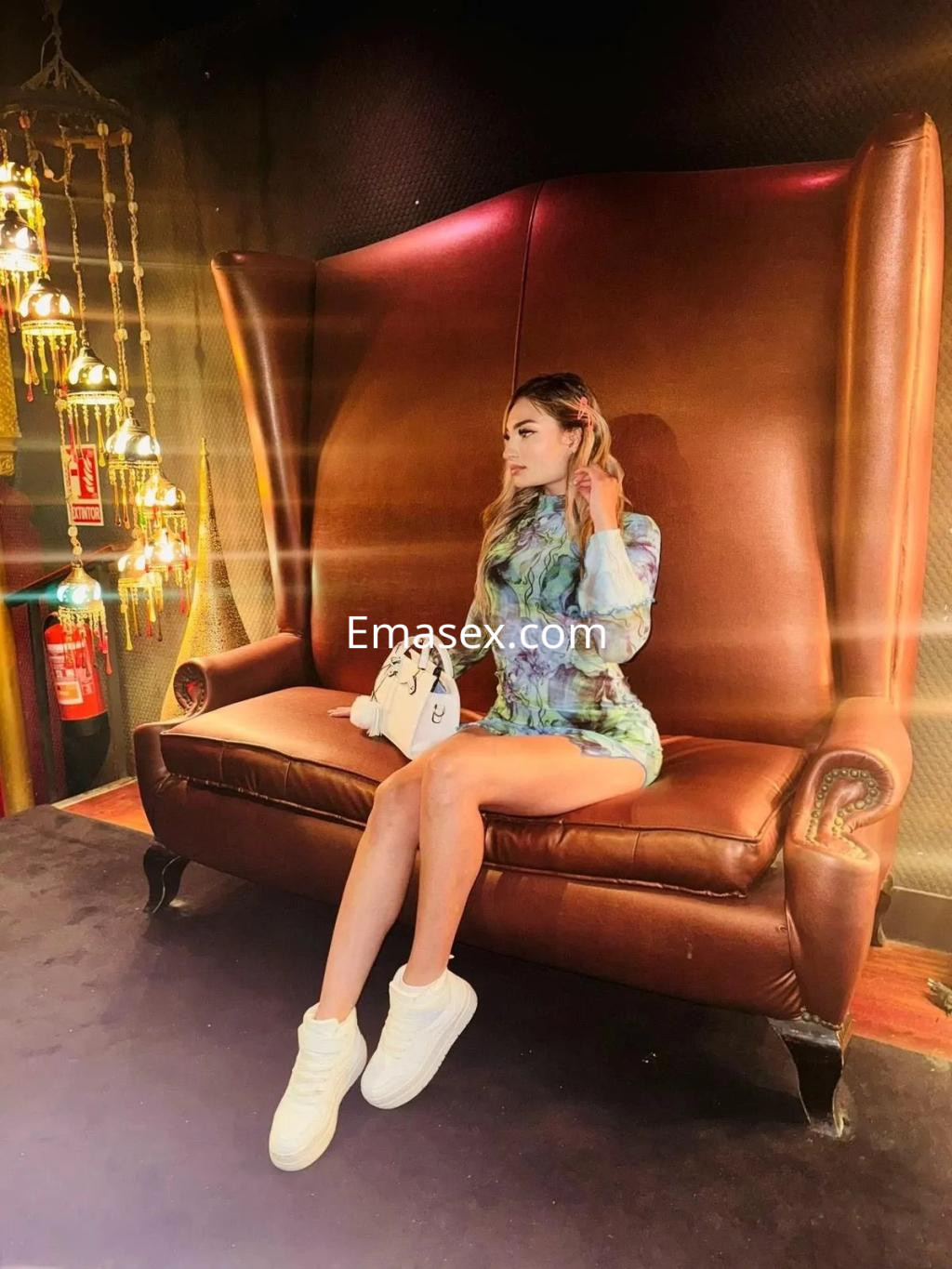 Escort en Valencia, sentada en un sofá de cuero, vistiendo un vestido colorido y zapatillas blancas.