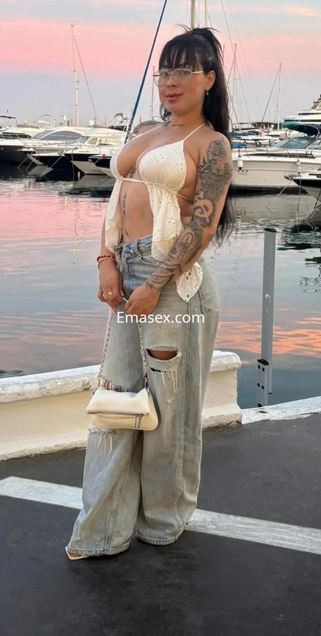 Escort en Valencia con blusa de crochet y jeans rasgados, posando frente al puerto al atardecer.