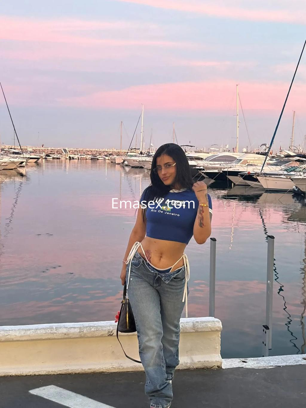 Escort en Valencia viste un top corto azul y jeans, posando junto al puerto con un atardecer colorido.