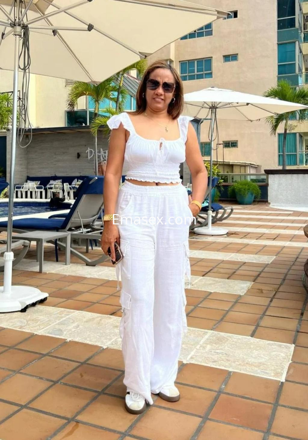 Puta en Madrid, luciendo un conjunto blanco de dos piezas con tirantes y pantalones largos, en un entorno de piscina.