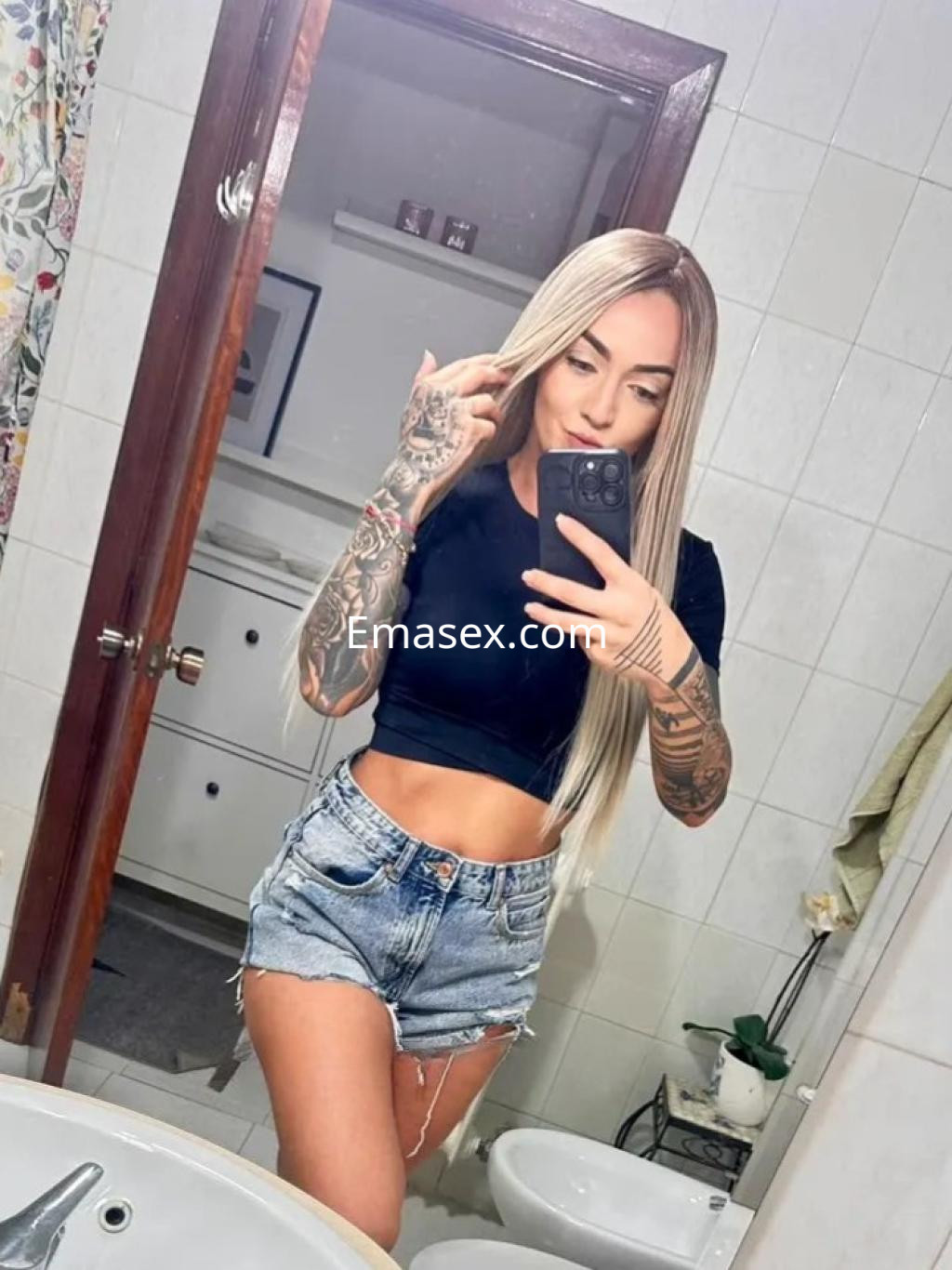 Puta en Valladolid con cabello largo y rubio, viste una camiseta negra y shorts de mezclilla, posando en el baño.