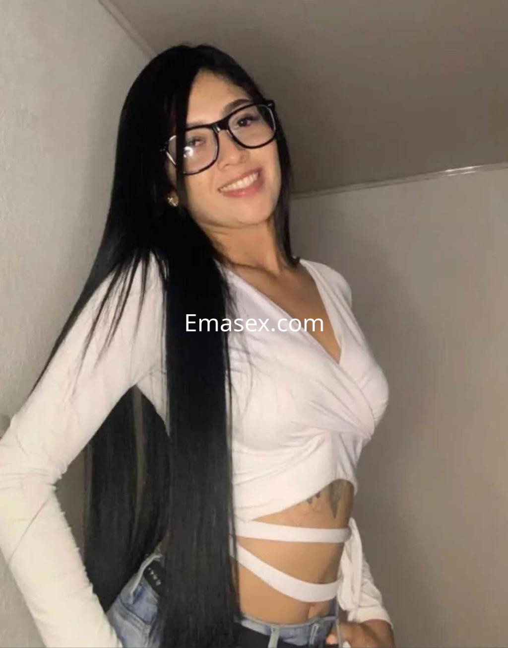 Escort en Alicante, con cabello largo y lacio, usa una blusa blanca ajustada y jeans, sonriendo en un ambiente interior.