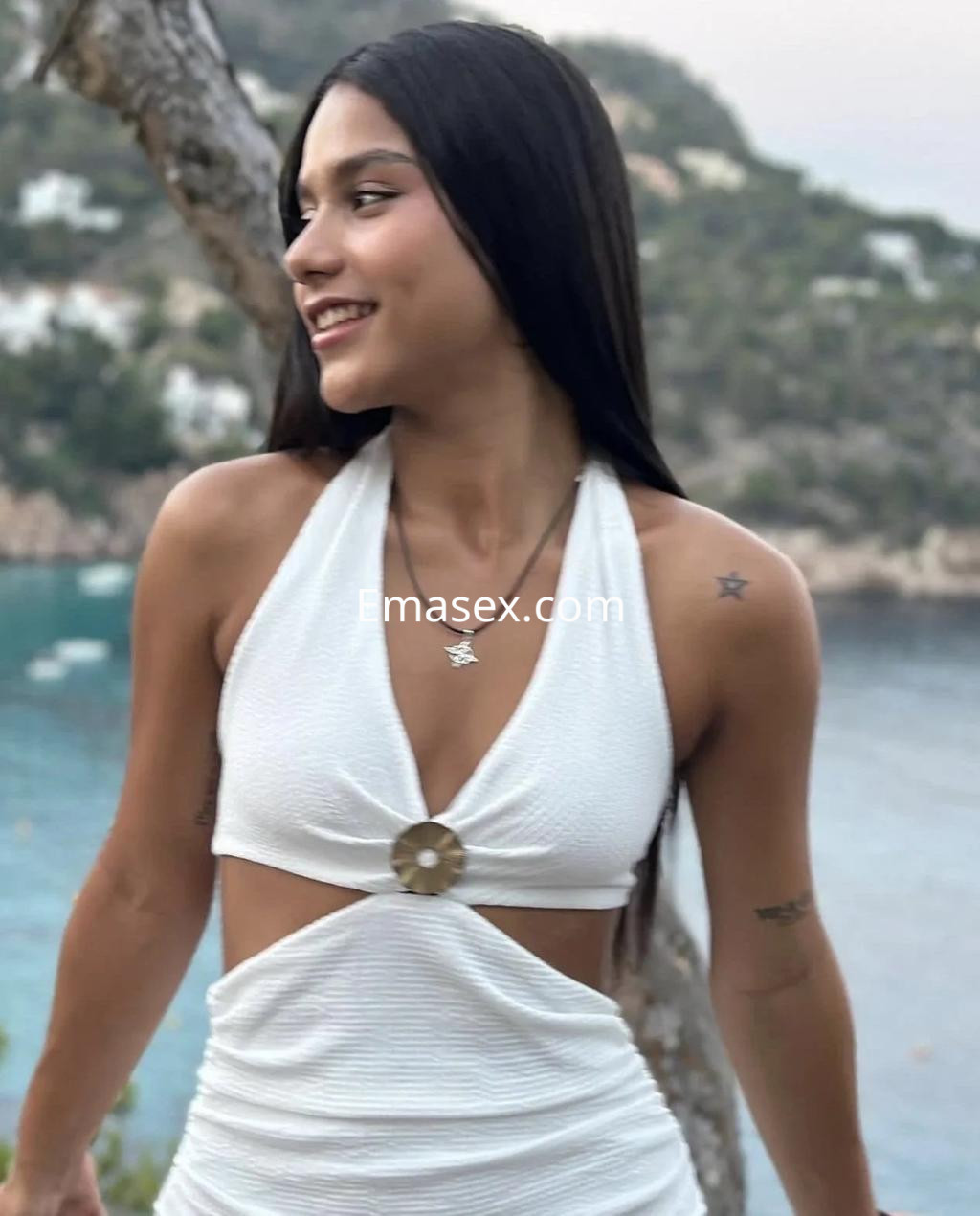 La escort en Alicante lleva un vestido blanco con detalle en el abdomen, sonriendo frente a un paisaje costero.