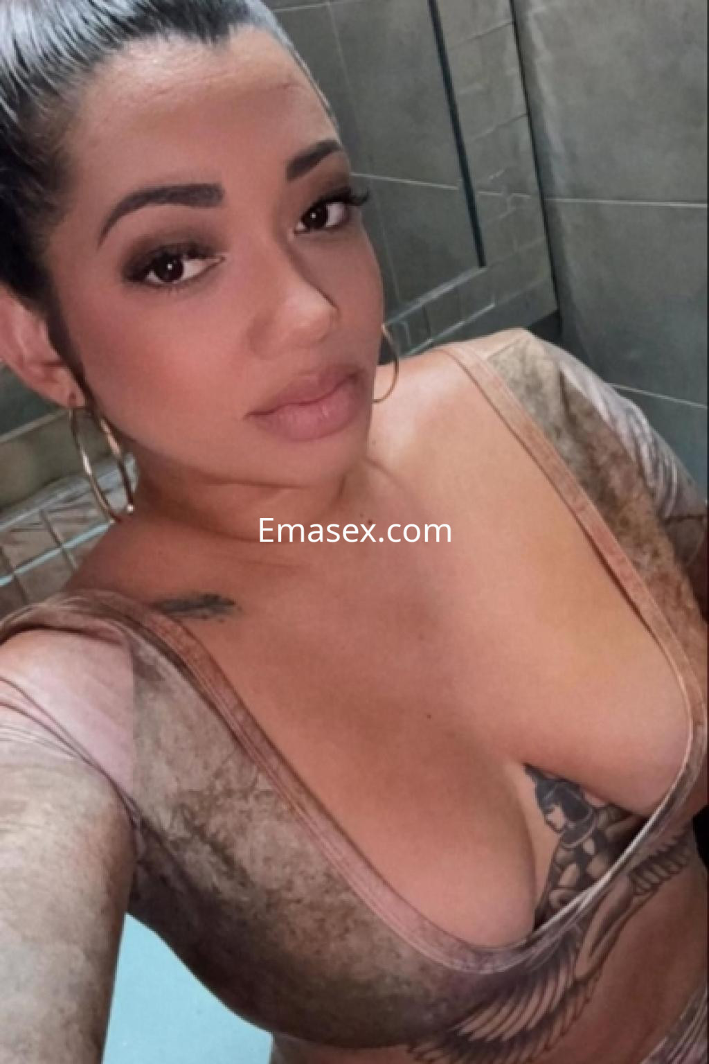 Escort en Almería con un top ajustado, pendientes grandes y un tatuaje visible en el pecho, posando en un baño.