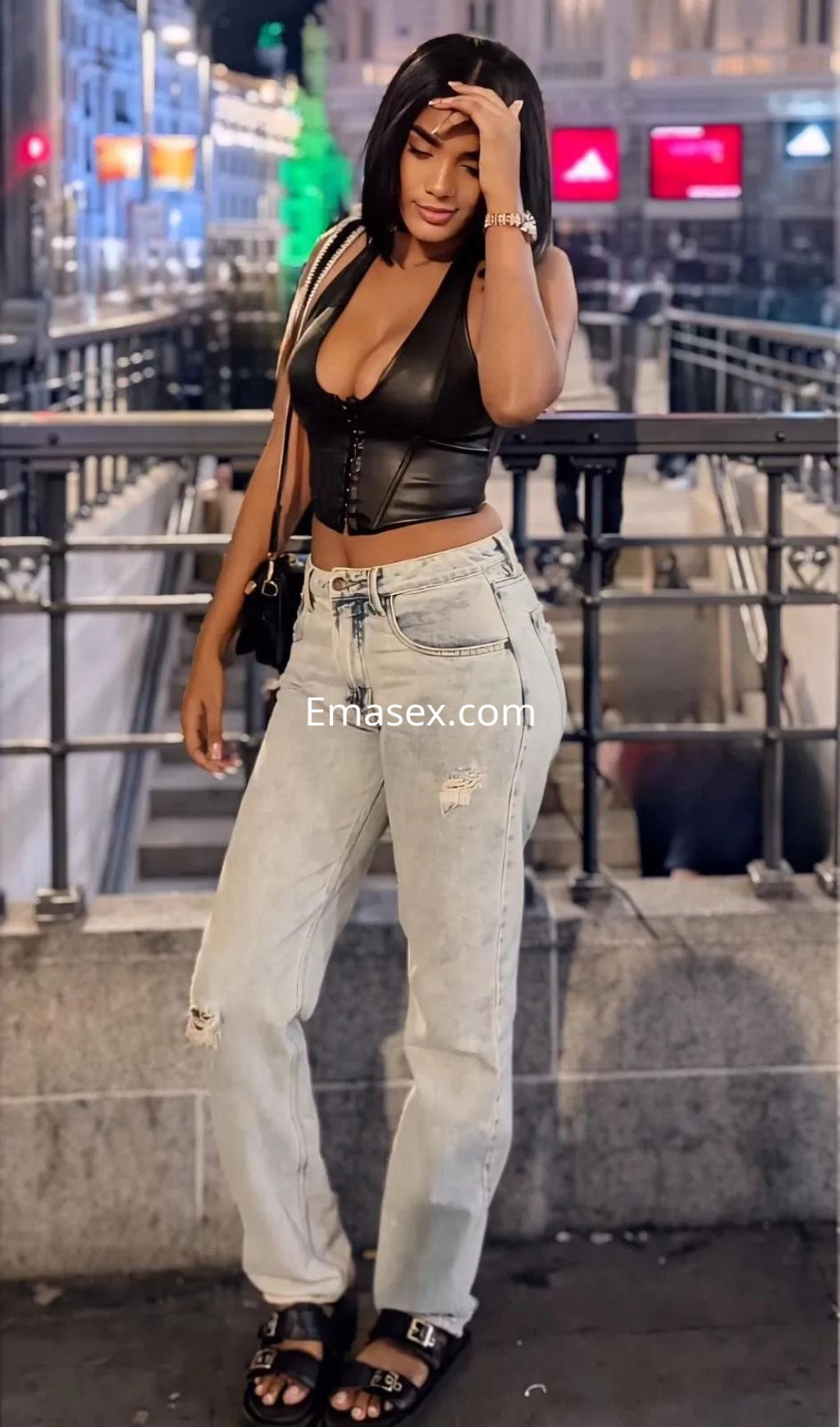 Puta en Palma de Mallorca con blusa de cuero negra y jeans claros, posando en un entorno urbano nocturno.