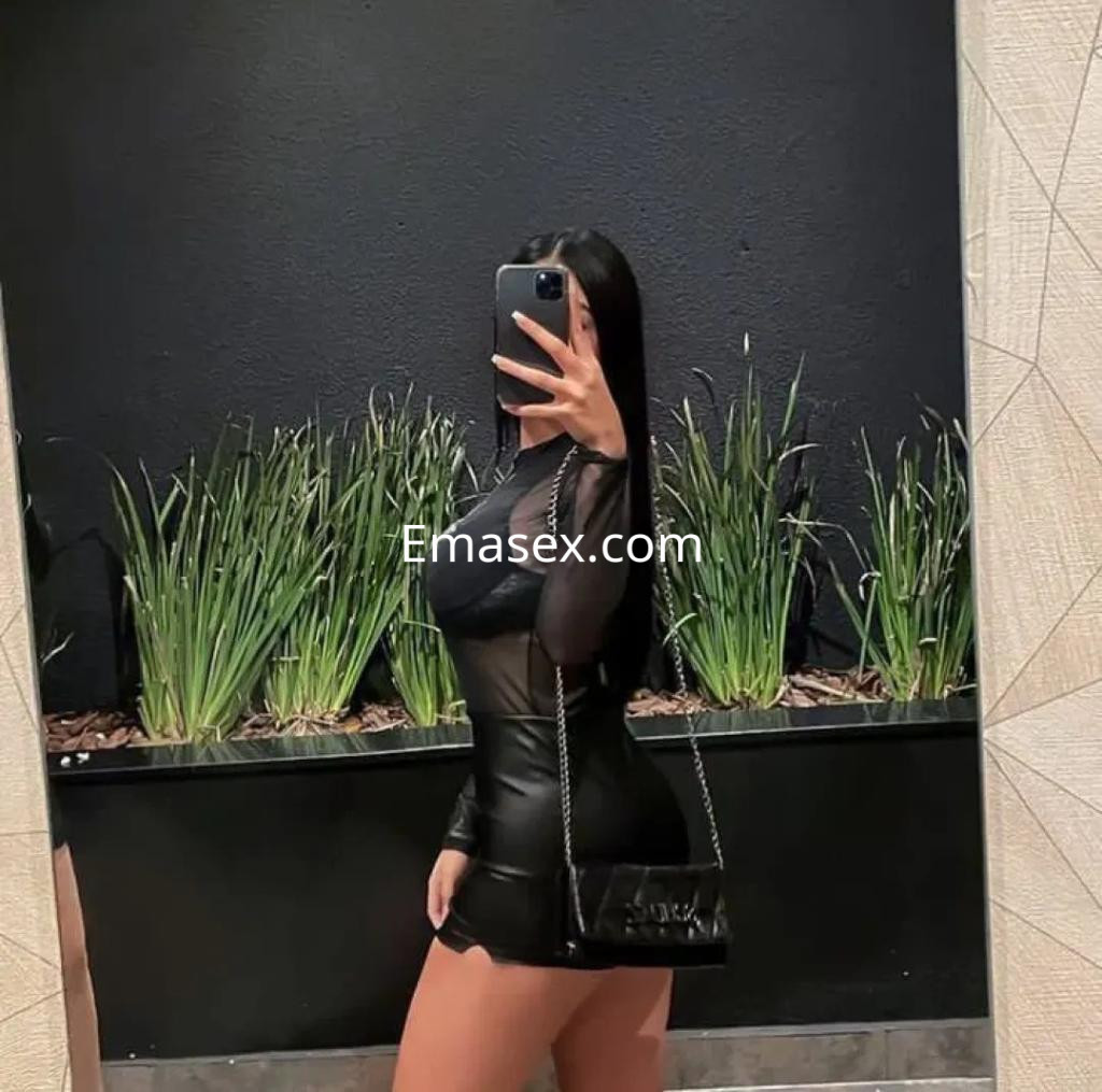 Escort en Cádiz posando frente a un espejo, con un vestido negro ajustado y una bolsa de mano.