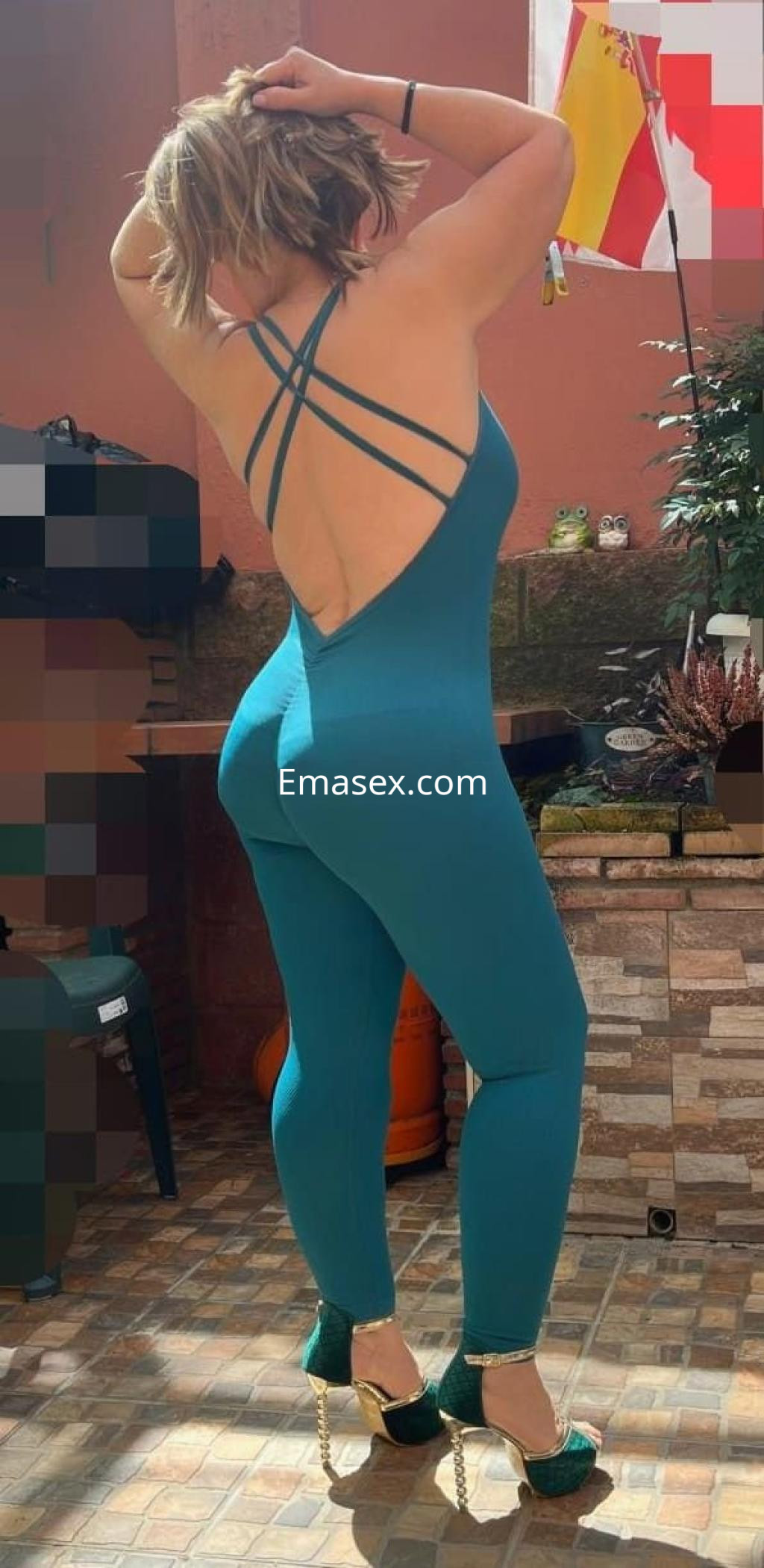 Escort en Cádiz posando de espaldas, vistiendo un mono azul con tirantes y tacones altos, en un ambiente exterior.