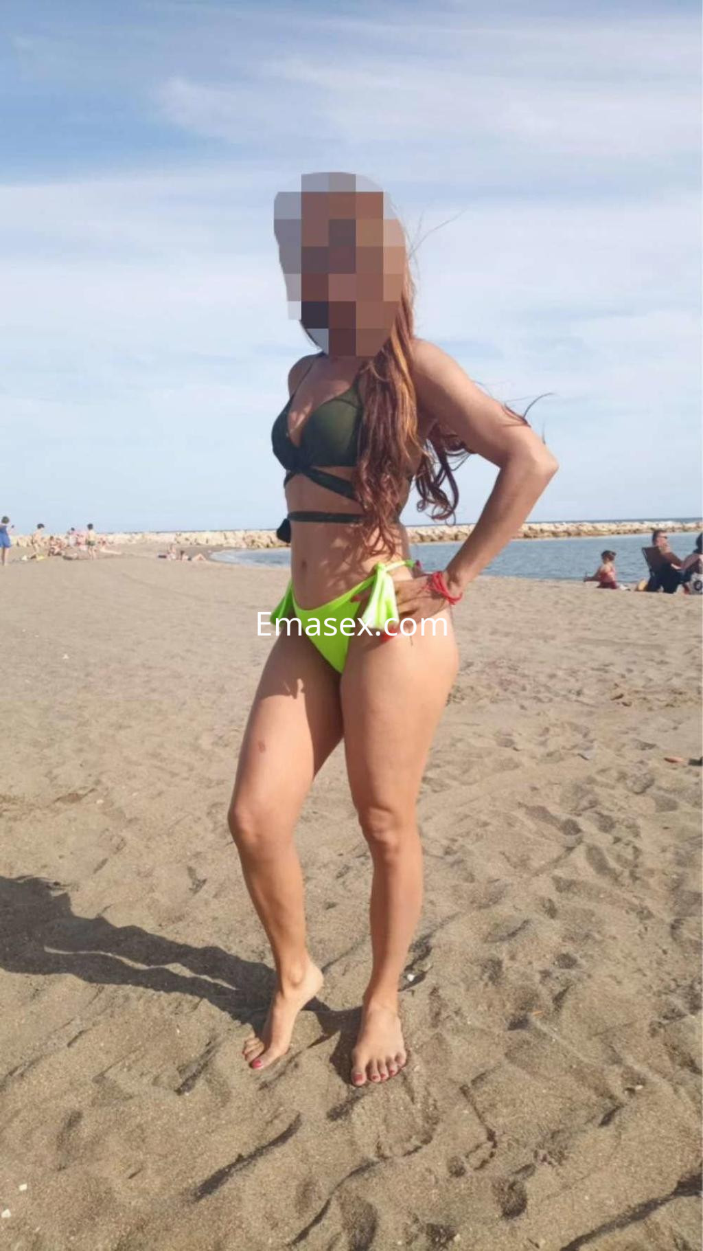 Escort en Cádiz, luciendo un bikini verde y una falda en la playa, con el mar de fondo y cabello largo.