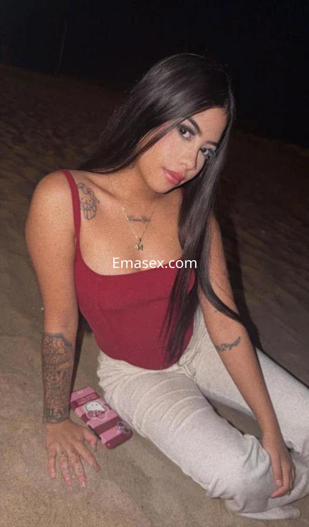 Escort en Castellón con top rojo y pantalones claros, sentada en la arena con el cabello largo y lacio.
