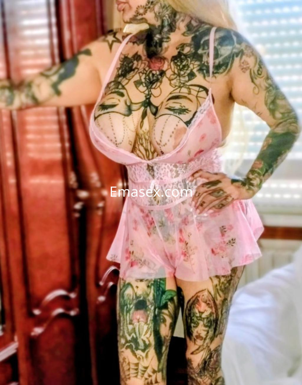 Escort en Ourense lleva un vestido lencero rosa claro con estampados florales y un cuerpo decorado con tatuajes.