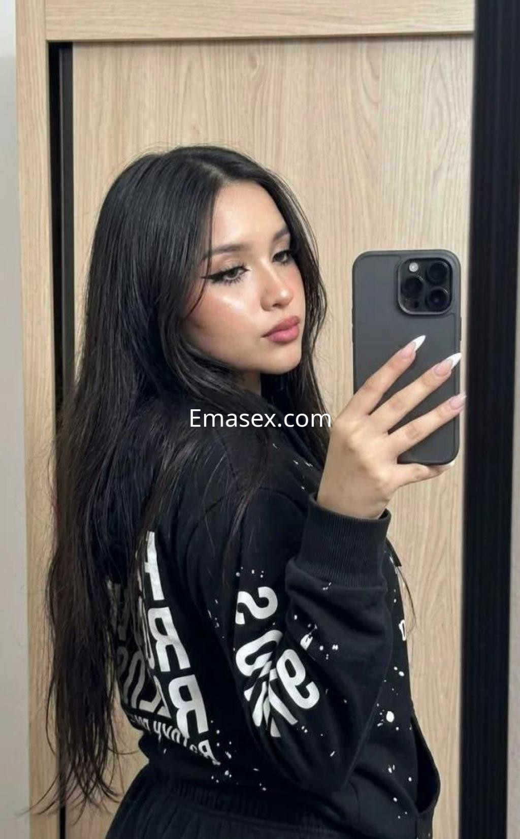 Escort en Oviedo con cabello largo y oscuro, luciendo un conjunto deportivo negro mientras sonríe y se toma una selfie.