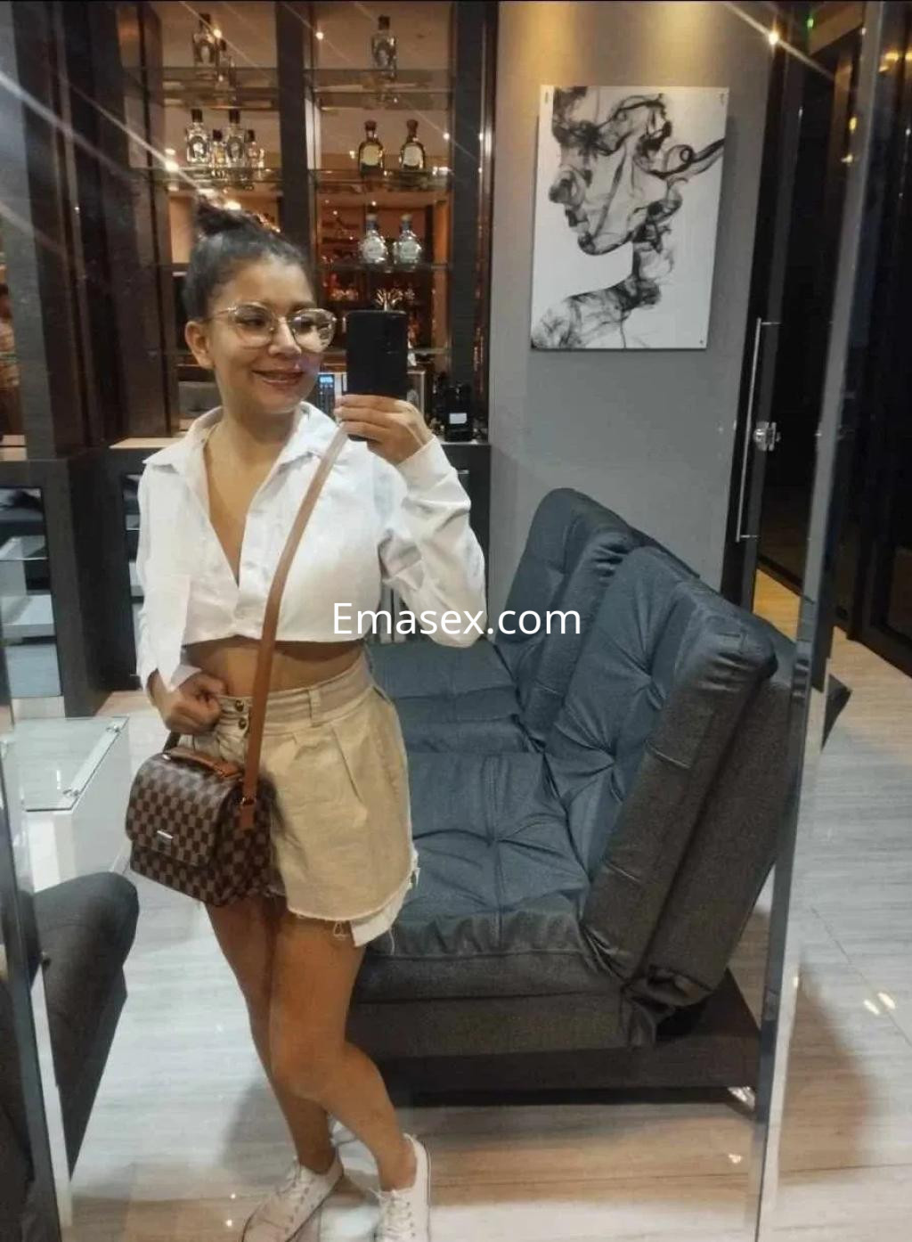 Escort en Oviedo con blusa blanca recortada y falda corta, posando en un moderno espacio interior.