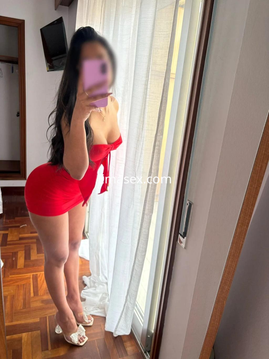 Escort en Vigo, posando frente a una ventana con un vestido rojo que resalta su figura, de cabello largo y oscuro.