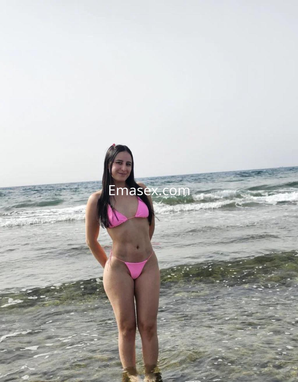 Puta en Tenerife vistiendo un bikini rosa en la playa, de pie junto al agua y el mar de fondo.