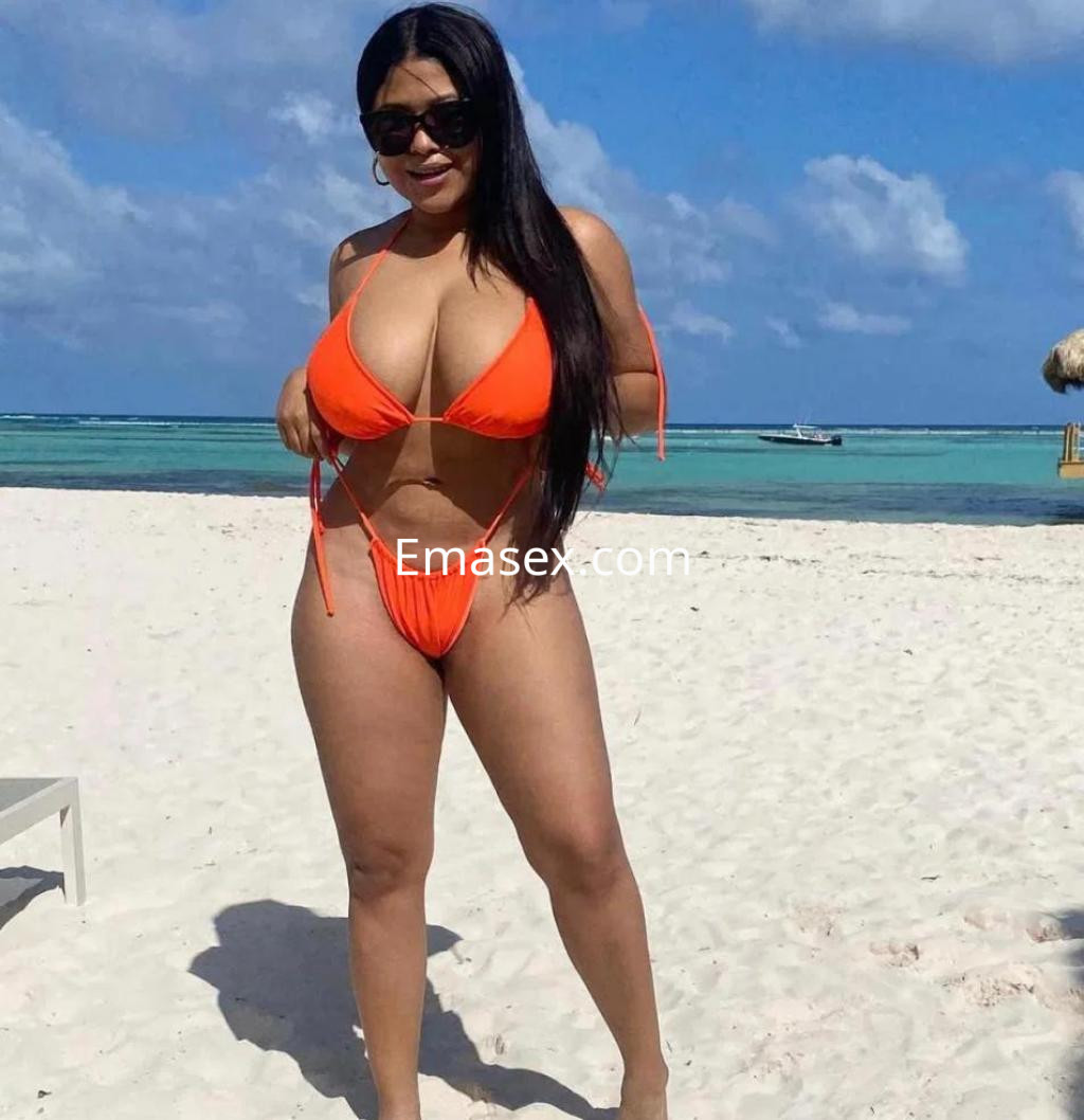 Escort en Tarragona con bikini naranja, posando en la playa con arena y mar de fondo, luciendo gafas de sol.