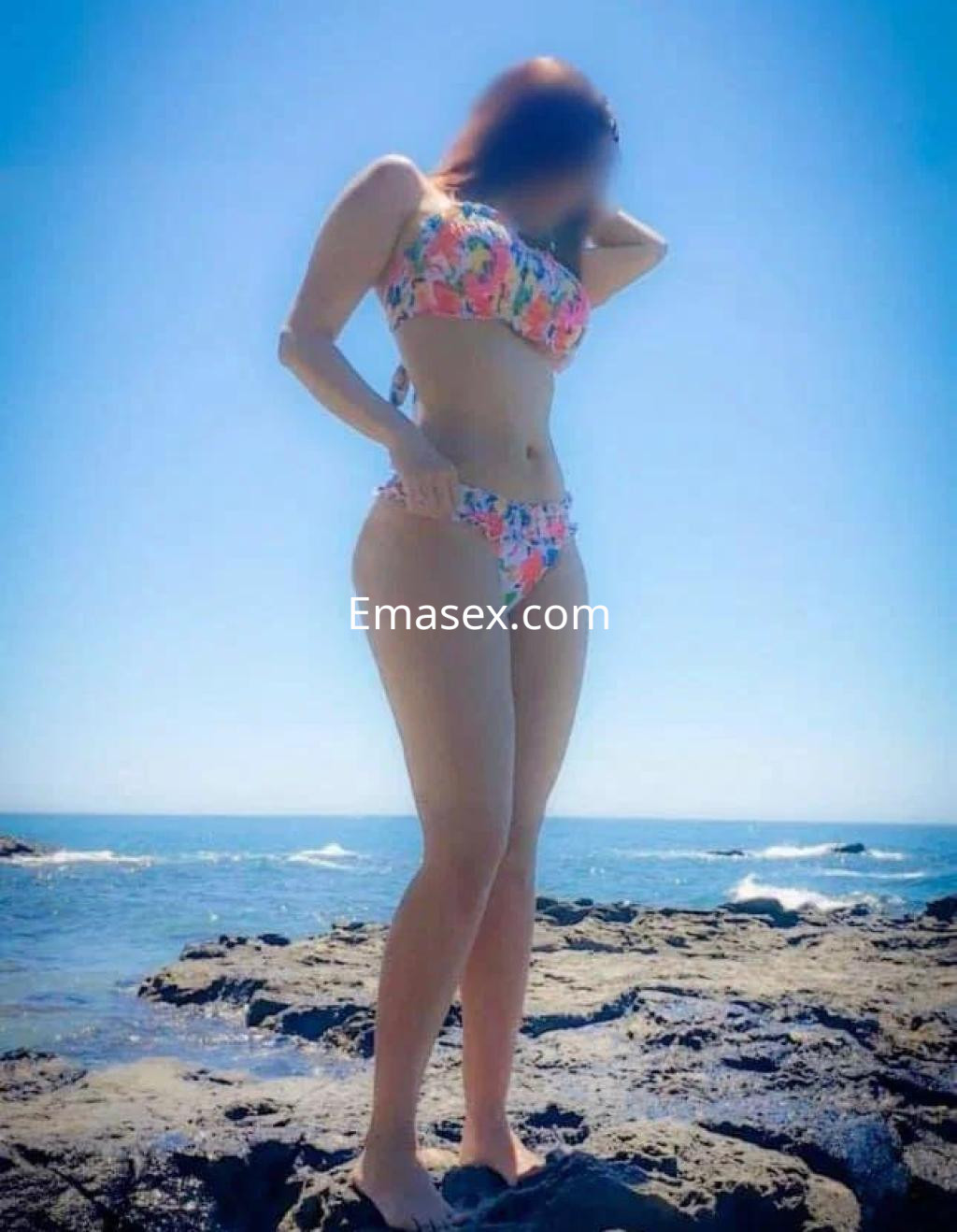Escort en Toledo con bikini colorido, posando en la playa con fondo de mar y rocas, bajo un cielo despejado.