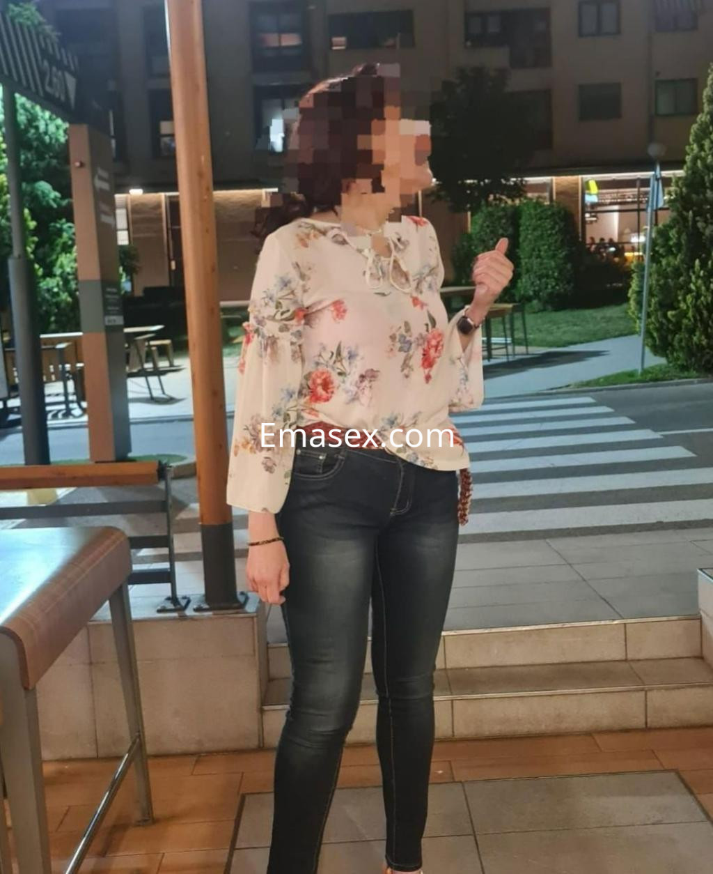 Escort en Madrid, luciendo una blusa de flores y jeans ajustados, posando al aire libre en un entorno urbano.