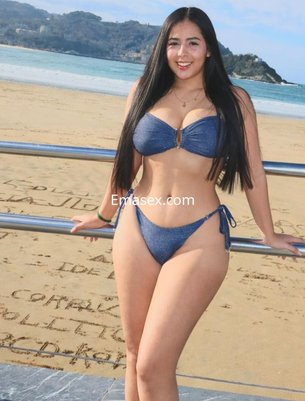 Escort en Vitoria luce un bikini azul con detalles en el centro, posando sonriente frente a la playa.