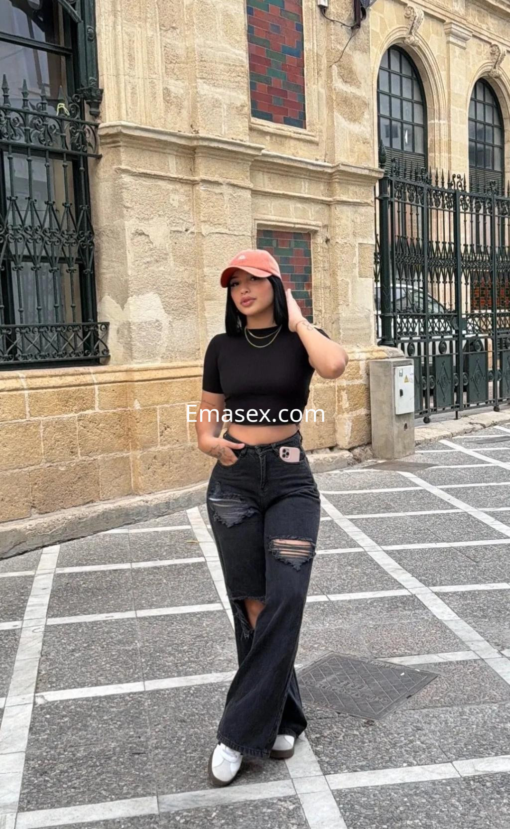 Puta en Cádiz con camiseta negra cropped, pantalones anchos rasgados y gorra, caminando por la calle.