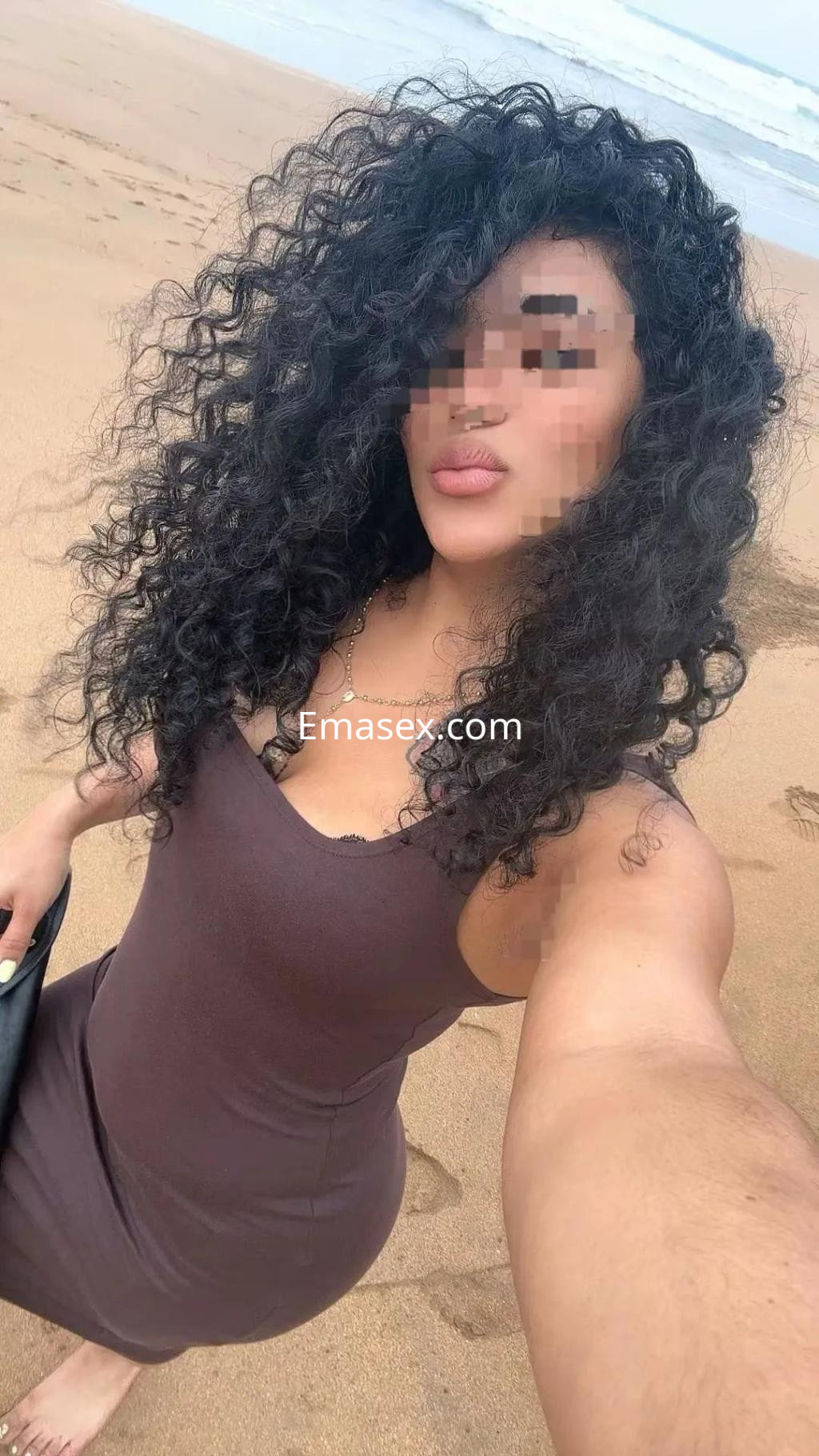 Puta con vestido ajustado marrón, cabello rizado y suelto, posando en la playa de Albacete.