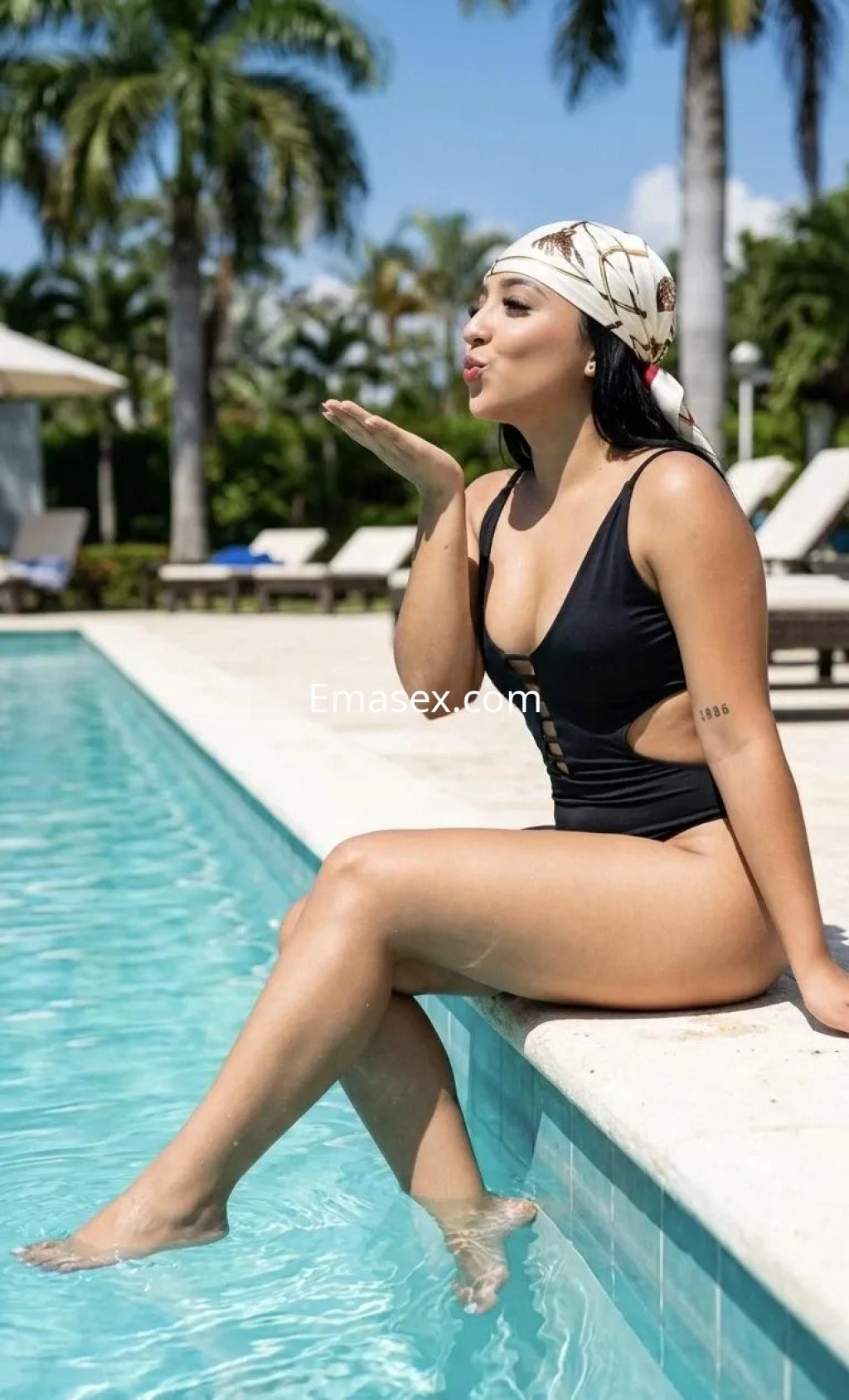 Puta en Burgos luciendo un traje de baño negro, sentada al borde de la piscina y sonriendo mientras lanza un beso.