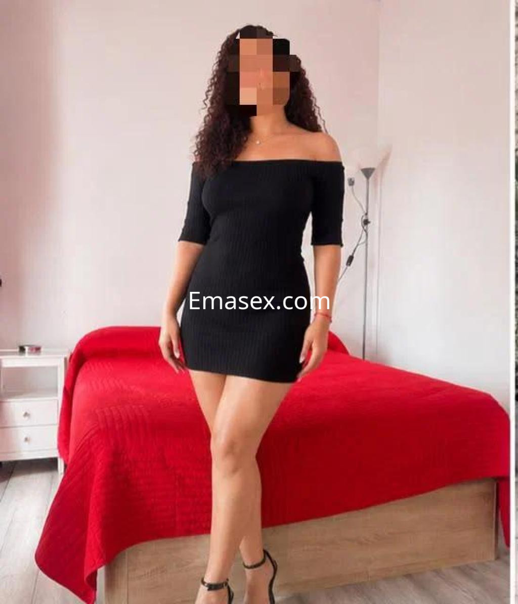 Escort en Cádiz con vestido negro ajustado, de pie en una habitación con una cama roja de fondo.