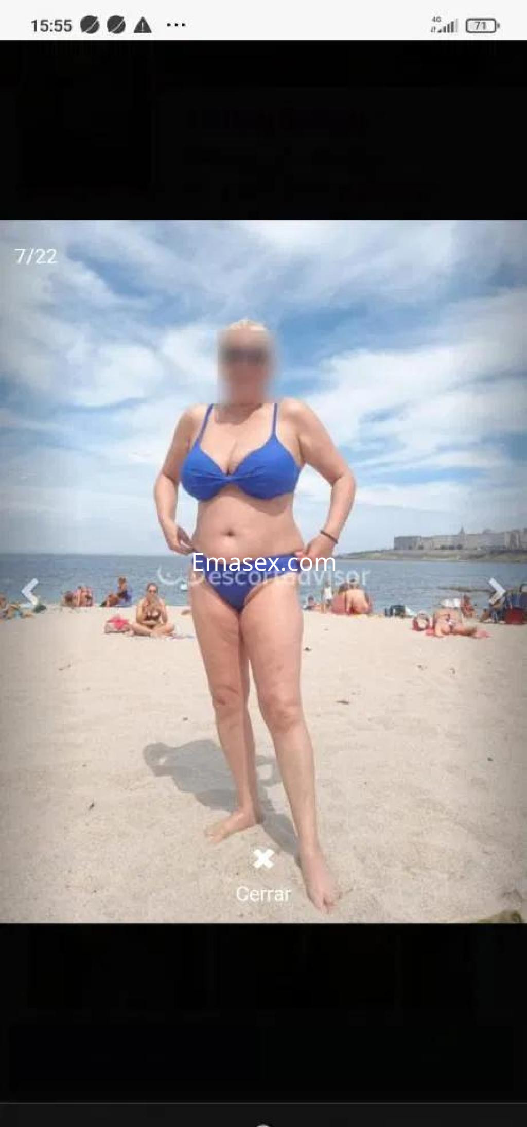 Escort en Coruña, de pie en la playa con un bikini azul, mostrando una pose relajada bajo un cielo nublado.