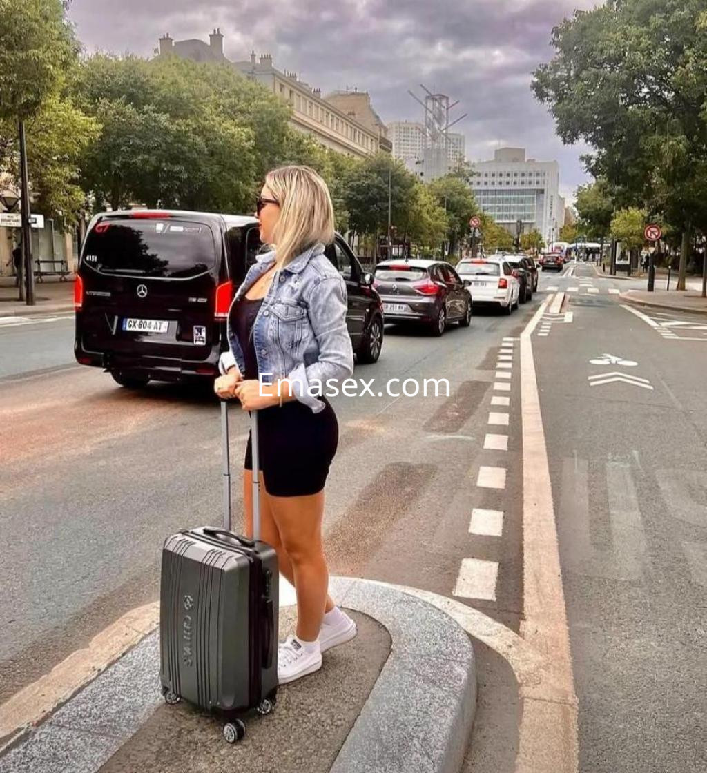 Puta de Coruña con cabello rubio y corto, viste una chaqueta de mezclilla y falda negra, esperando en la calle.
