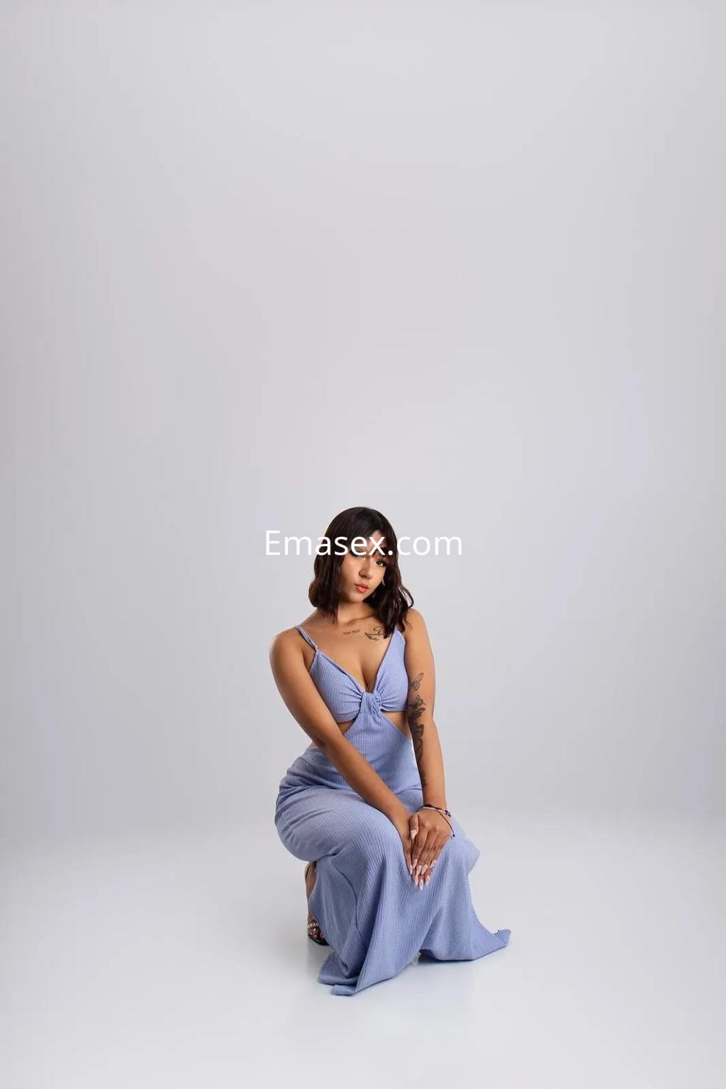 Escort sentada en una pose elegante, vistiendo un vestido azul claro, con cabello oscuro y lacio, en fondo neutro en Granada.