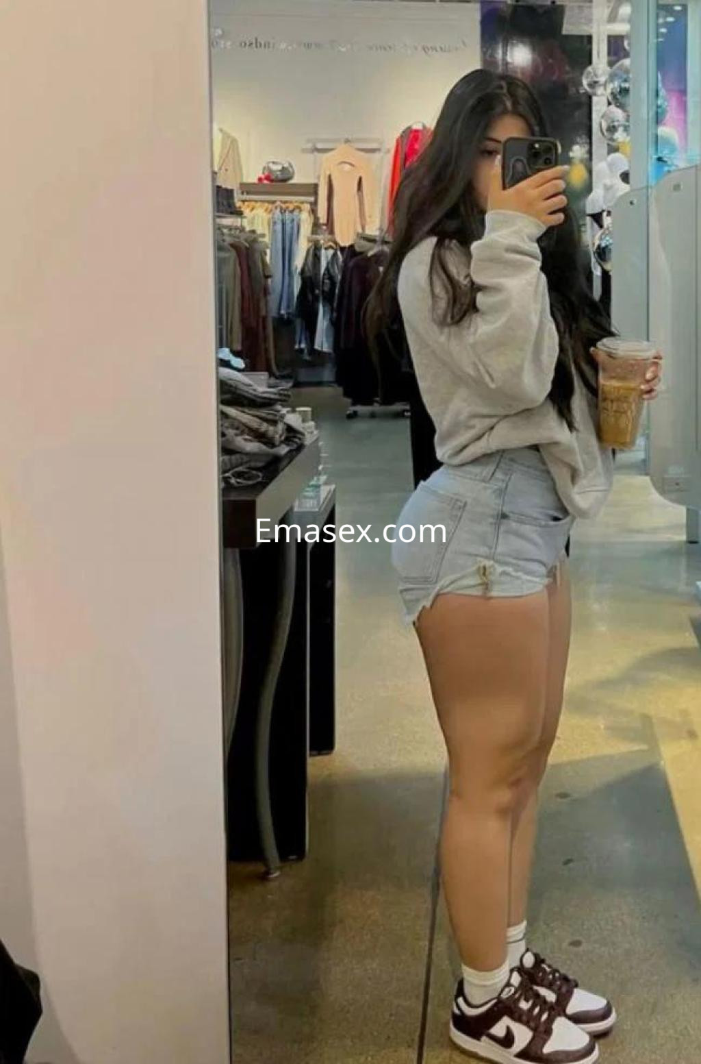 Escort en Huelva con cabello largo y oscuro, viste sudadera gris y shorts de mezclilla, sosteniendo un café.