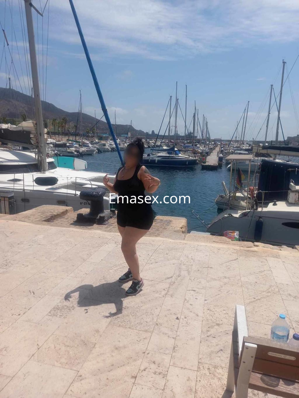 Escort en Málaga posando en un puerto deportivo, luciendo un vestido negro y zapatillas deportivas.