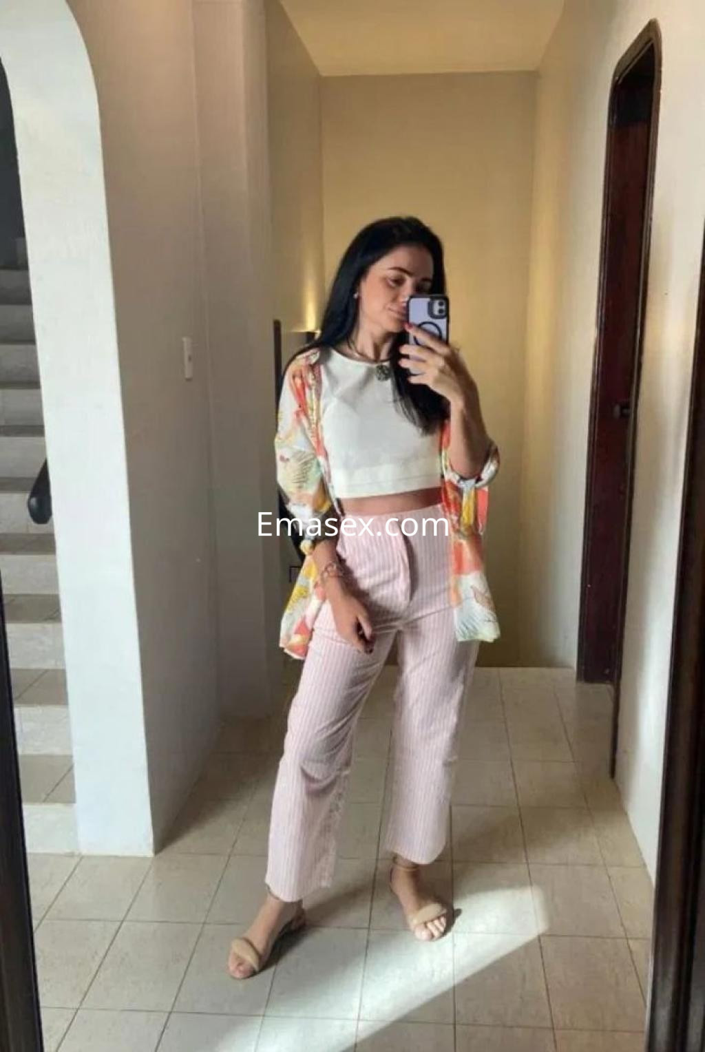 Puta en Málaga posa en un espejo, vistiendo un top blanco y pantalones de rayas, con una chaqueta floral.