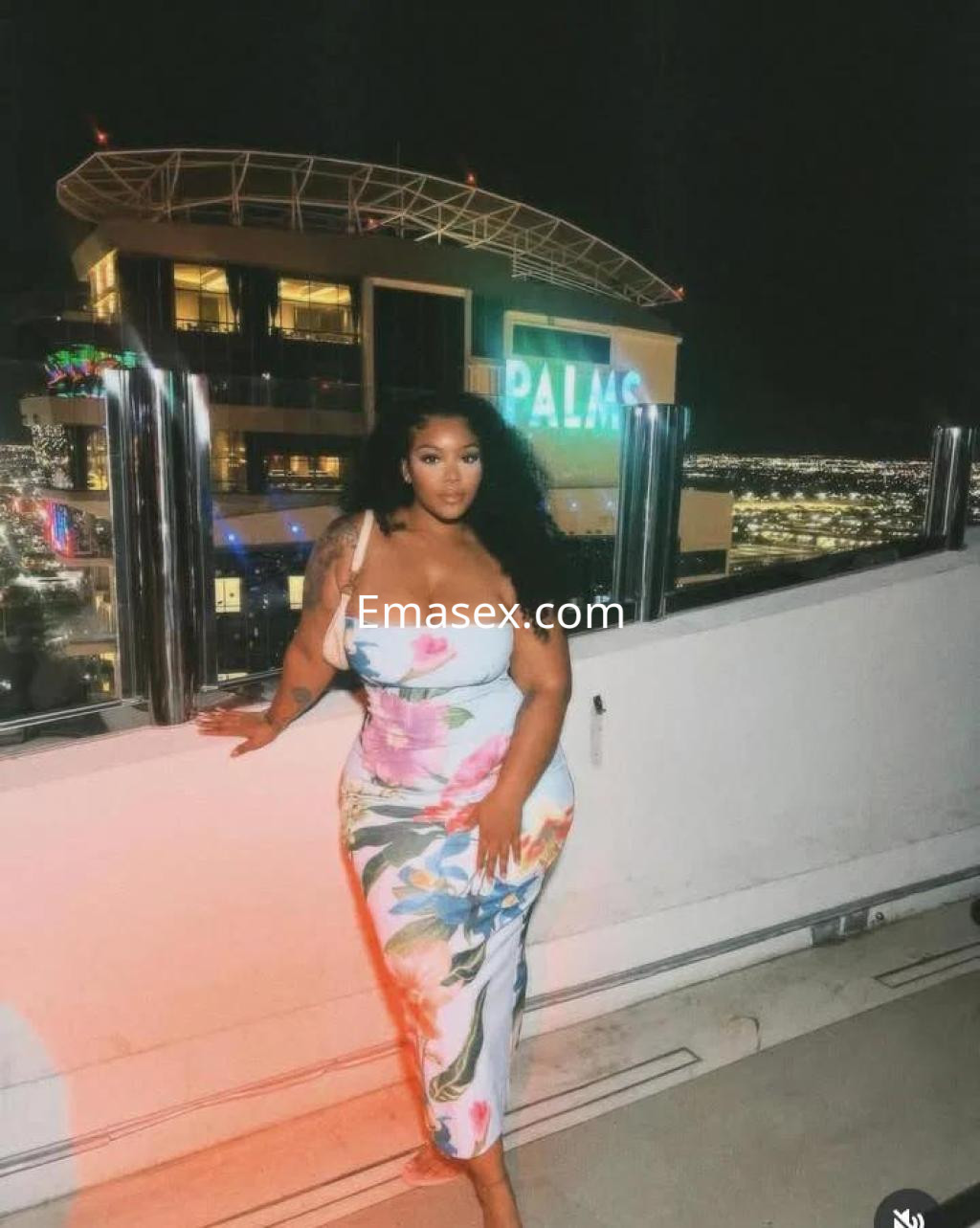 Escort en Murcia con vestido floral ajustado, posando en una terraza con vistas nocturnas.