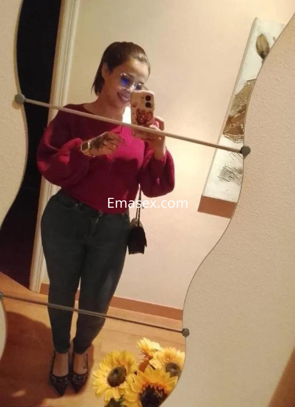 La escort en Murcia se toma una selfie frente a un espejo, luciendo un suéter rojo y jeans ajustados.
