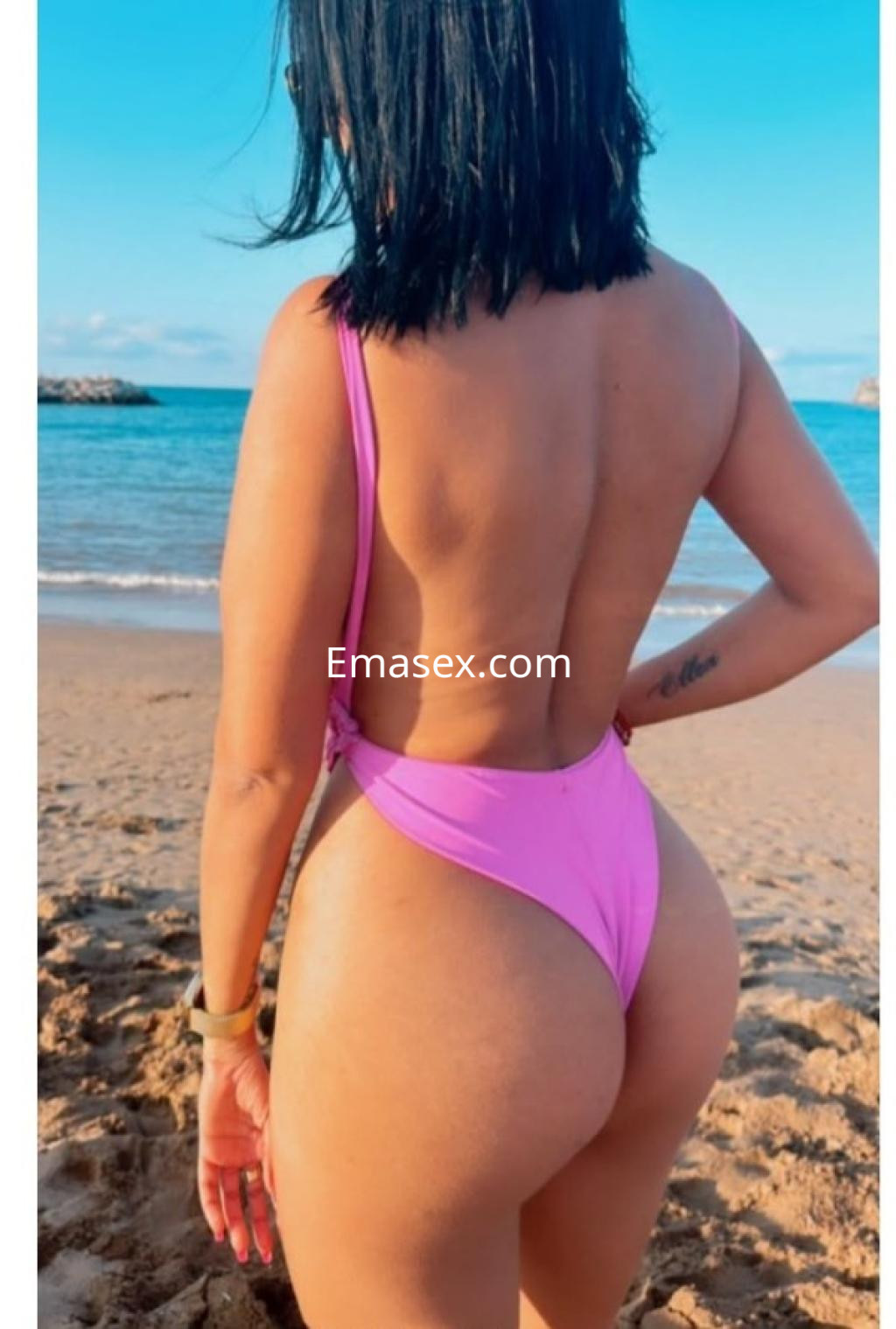 Escort en Ourense posando de espaldas en la playa, luciendo un traje de baño rosa y cabello corto.
