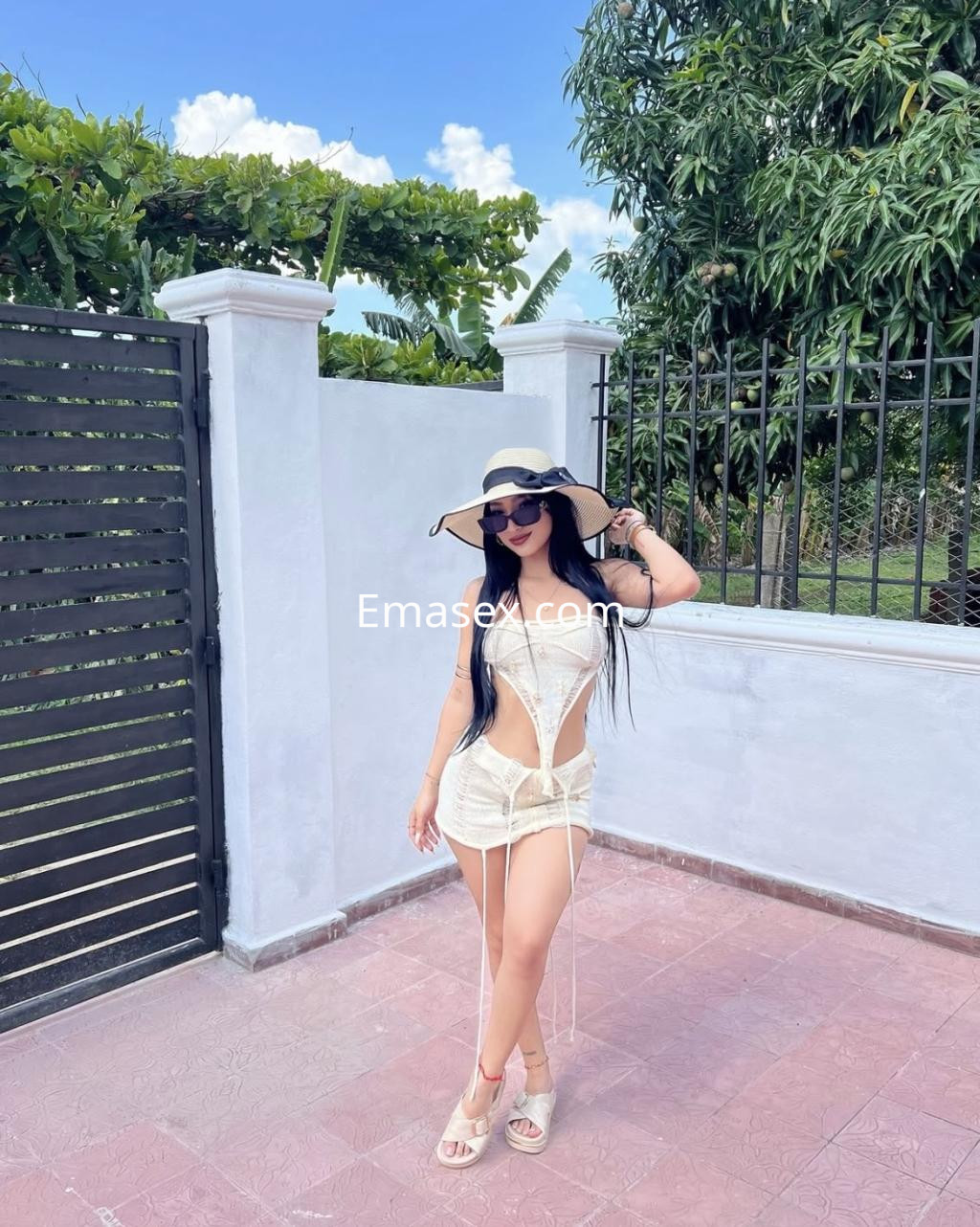 La puta luce un vestido blanco ajustado y un sombrero grande, posando en un entorno soleado en Santander.