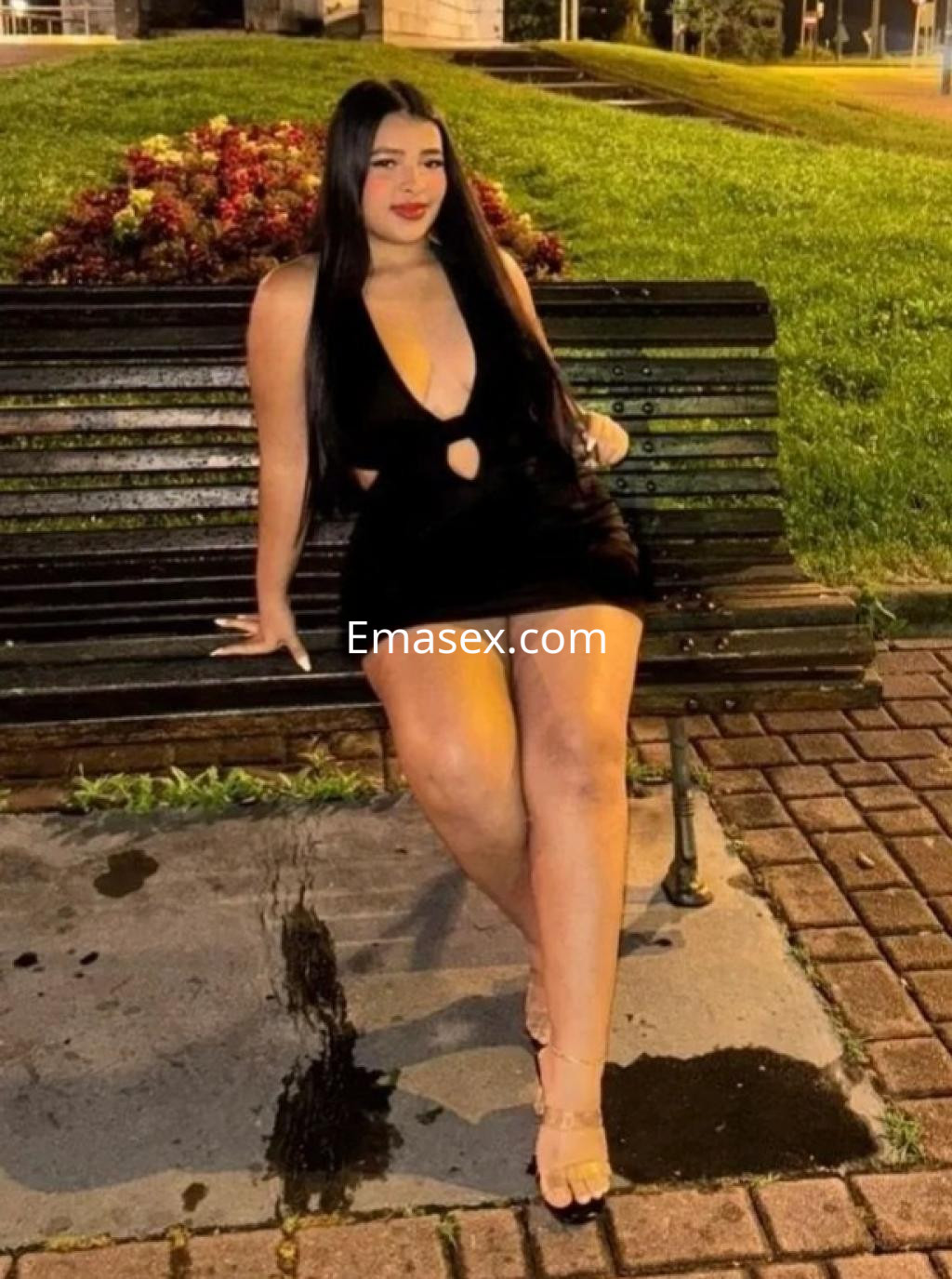 Escort en Bilbao sentada en un banco, luciendo un vestido negro con escote y sandalias, en un entorno urbano.