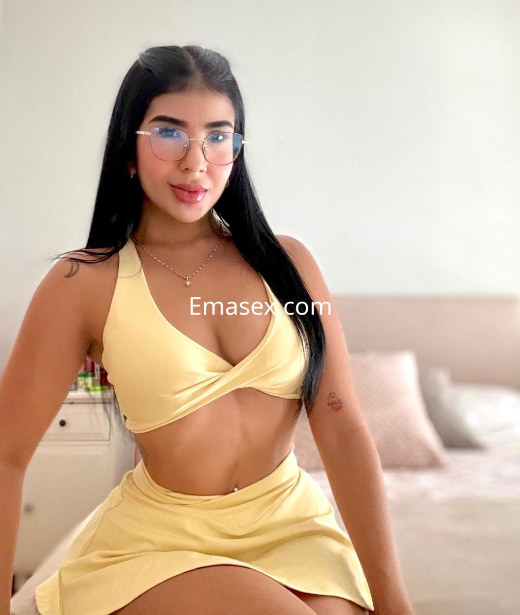 Puta en Pamplona luce un conjunto amarillo con top cruzado y falda, posando con gafas en un entorno luminoso.