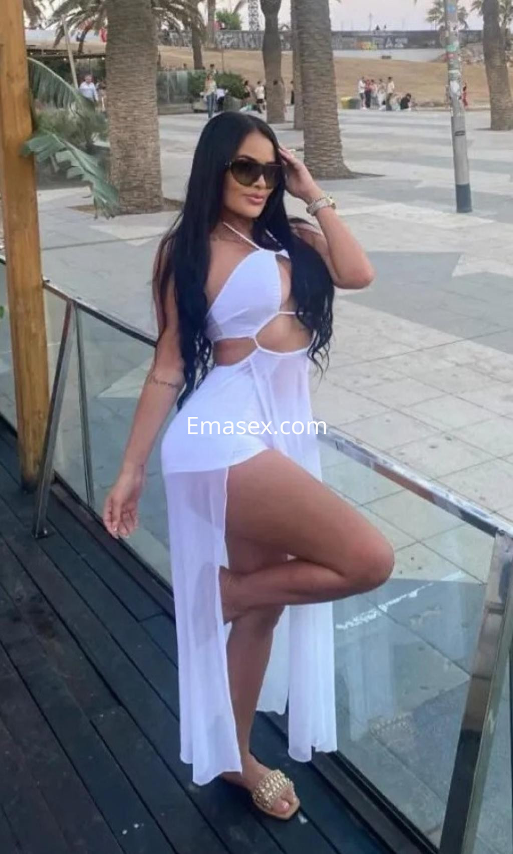 Puta de cabello largo y oscuro, viste un vestido blanco transparente, posando en Valencia junto a palmeras.