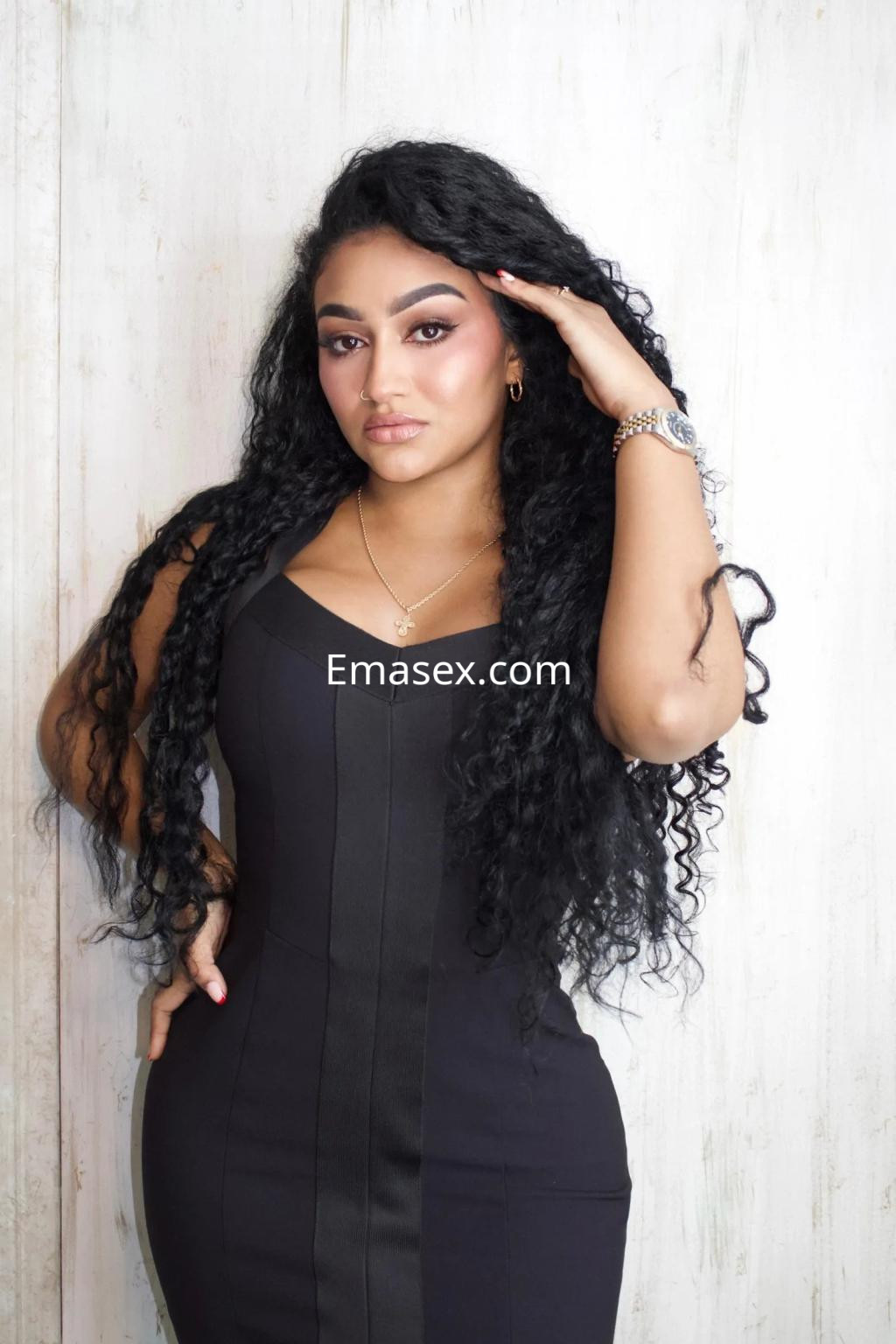Escort en Alicante con un vestido negro ajustado, cabello rizado y largo, posando con una mano en la frente.