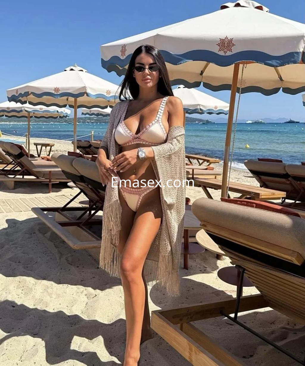 Escort en Alicante con bikini claro y un chal de flecos, posando junto a sombrillas en la playa.