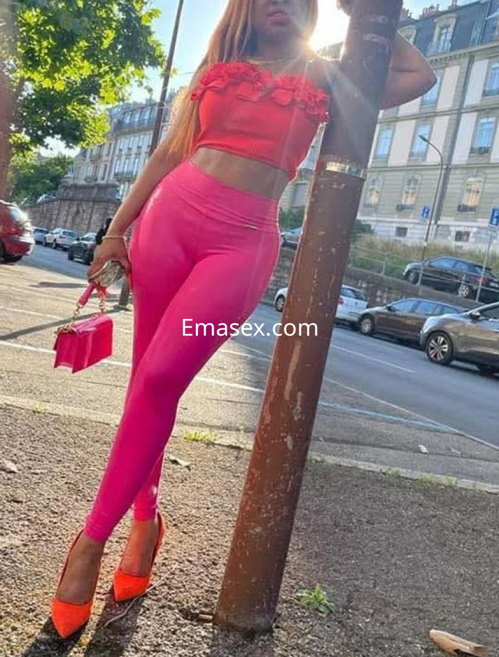 Escort en Coruña, luciendo un top rojo con detalles, leggins rosas y tacones altos, posando junto a un poste.