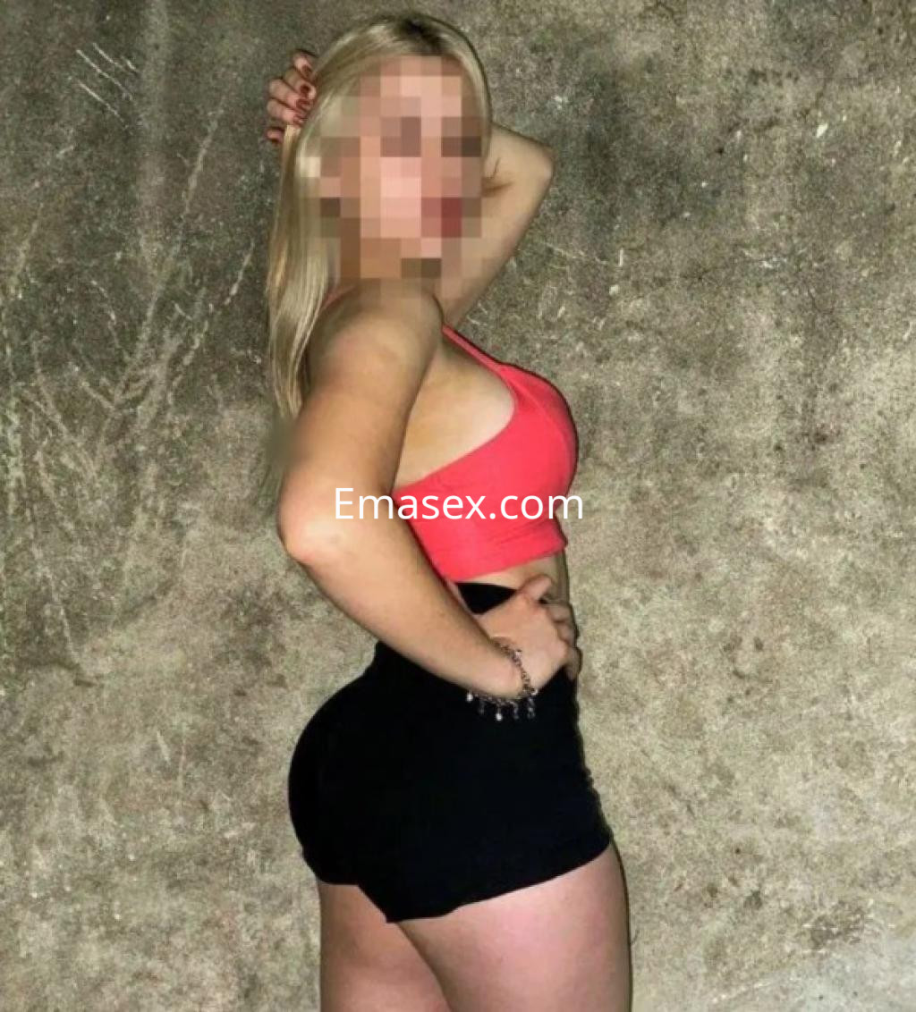 Puta con cabello rubio y lacio, viste un top rojo y shorts negros, posando de perfil en Vigo.