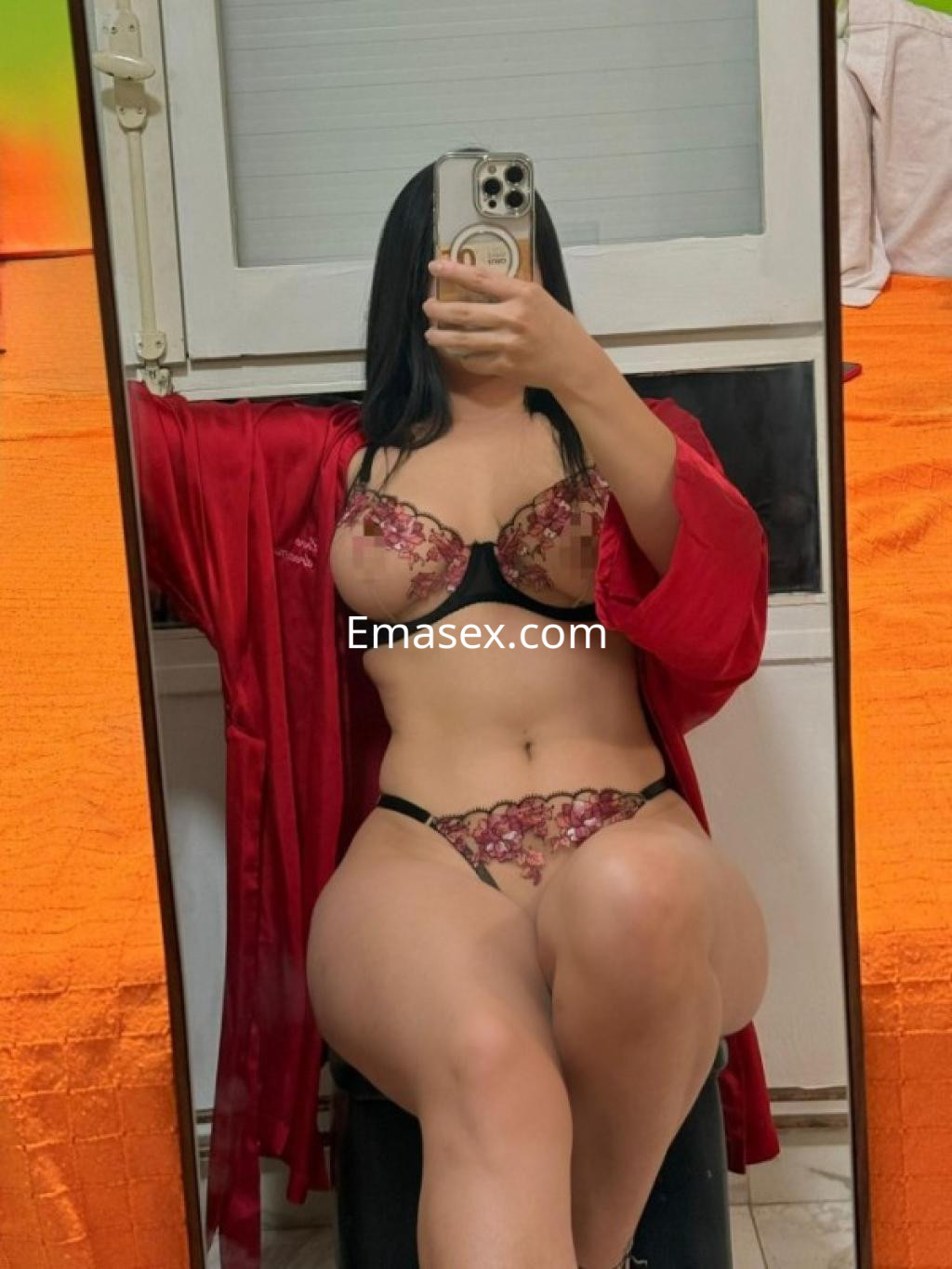 Escort en Salamanca posando frente a un espejo, vistiendo lencería negra con detalles florales y un robe rojo.