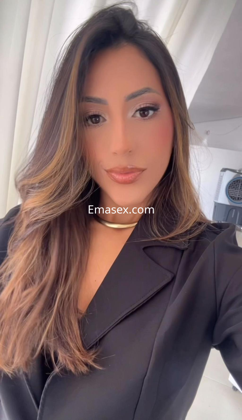 Escort en Vitoria con cabello largo y suelto, vistiendo un blazer negro y un collar dorado, posando de forma elegante.